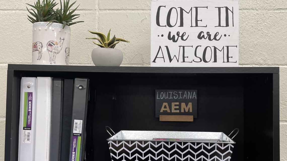 Shelf with plants and a sign reading "come in we are awesome and Louisiana AEM"