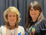 two women, woman on the left is blonde with a beige sweater, woman on the right brunette with black dress