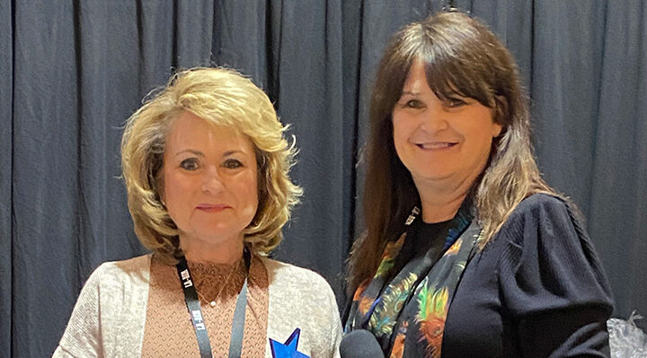 two women, woman on the left is blonde with a beige sweater, woman on the right brunette with black dress